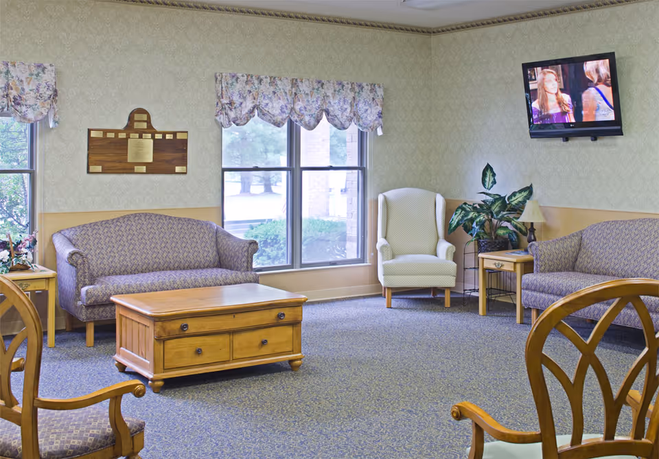 A cozy lounge with upholstered sofas and armchairs, a wooden coffee table, potted plant, wall-mounted TV and windows.
