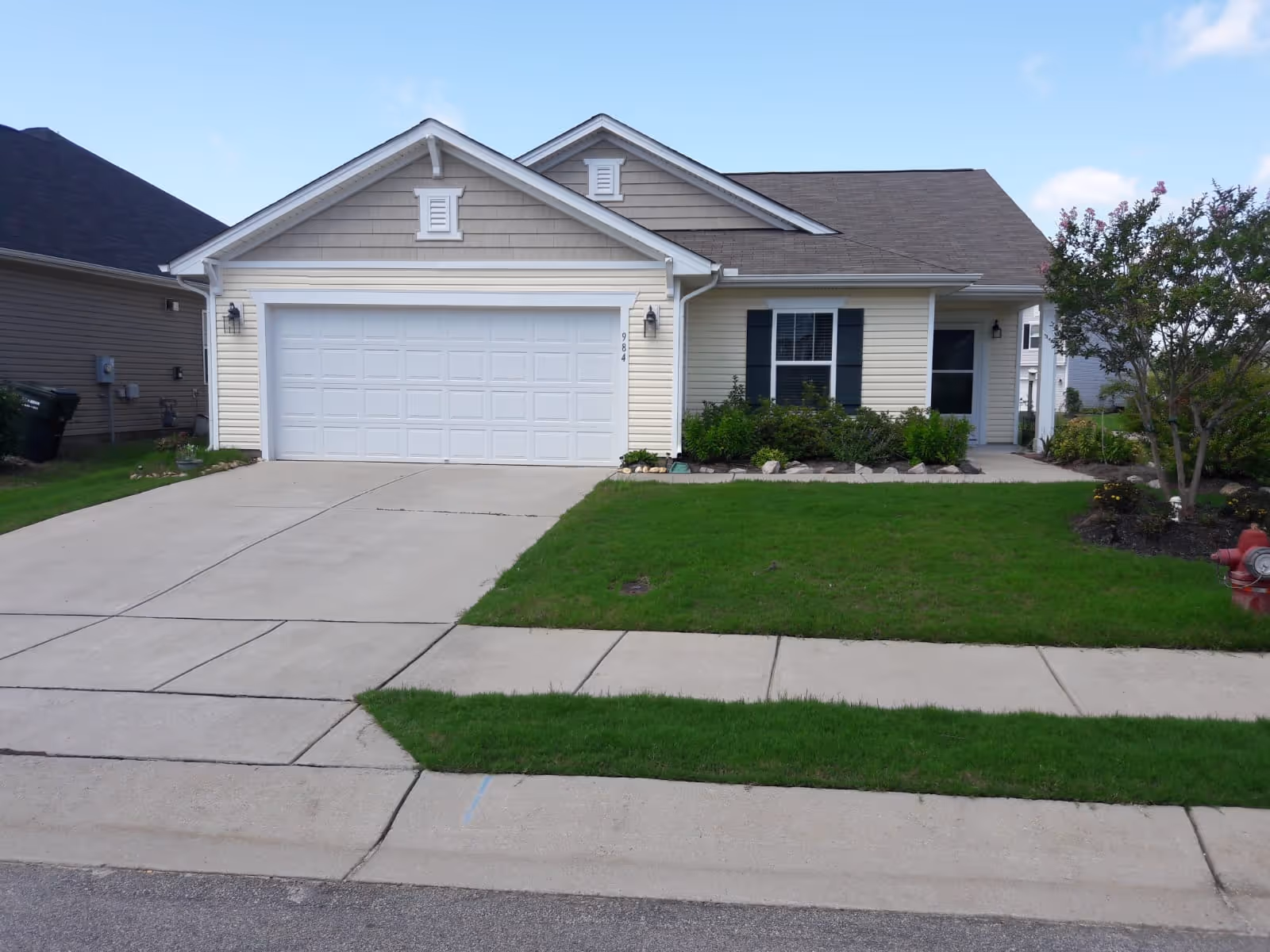 Front exterior view of a single-story house with a two-car garage, beige siding, a small front porch, green lawn, and a tree on the right side.