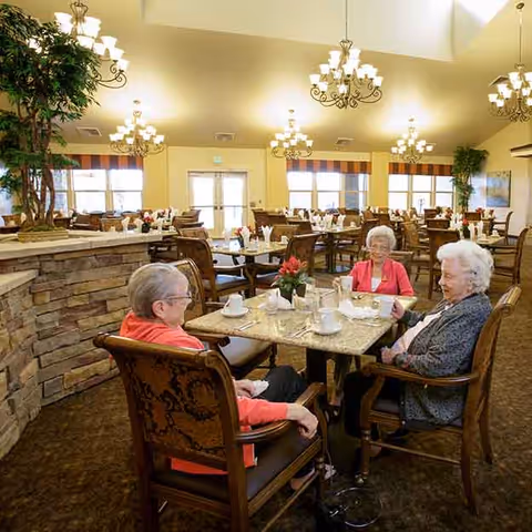 Three elderly women sitting around a table in a spacious, well-lit dining room with multiple tables and chairs. The room features large windows with striped valances, chandeliers hanging from the ceiling, and decorative plants. The table is set with cups, saucers, and a small flower arrangement.