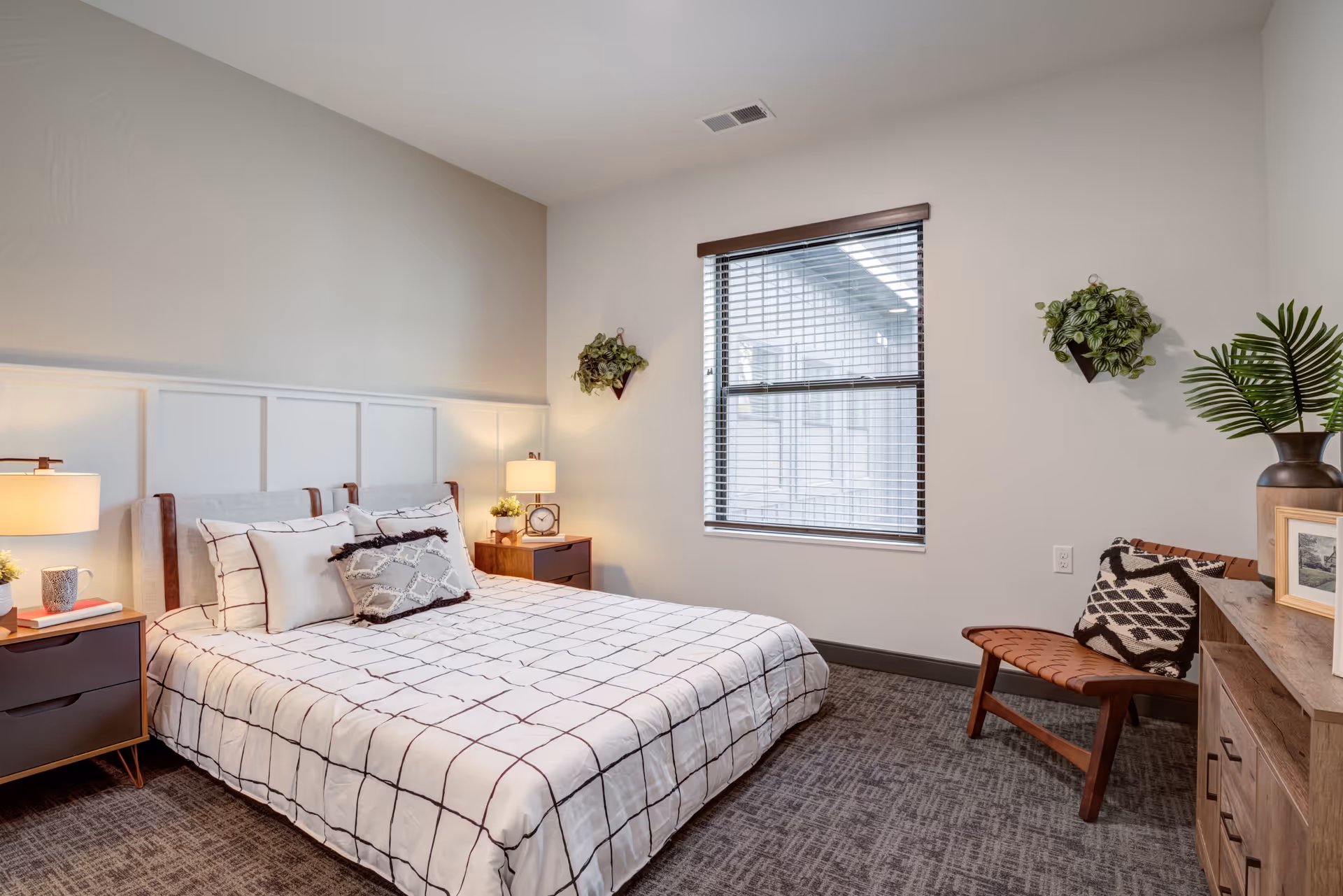 A neatly arranged bedroom with a bed featuring a white and black checkered bedspread and multiple pillows. There are two wooden nightstands on either side of the bed, each with a lamp and decorative items. A window with blinds is centered on the far wall, flanked by two small wall-mounted plants. To the right, there is a wooden dresser with a large vase containing green leaves and a framed picture, and a wooden chair with a patterned cushion. The room has neutral-colored walls and carpeted flooring.