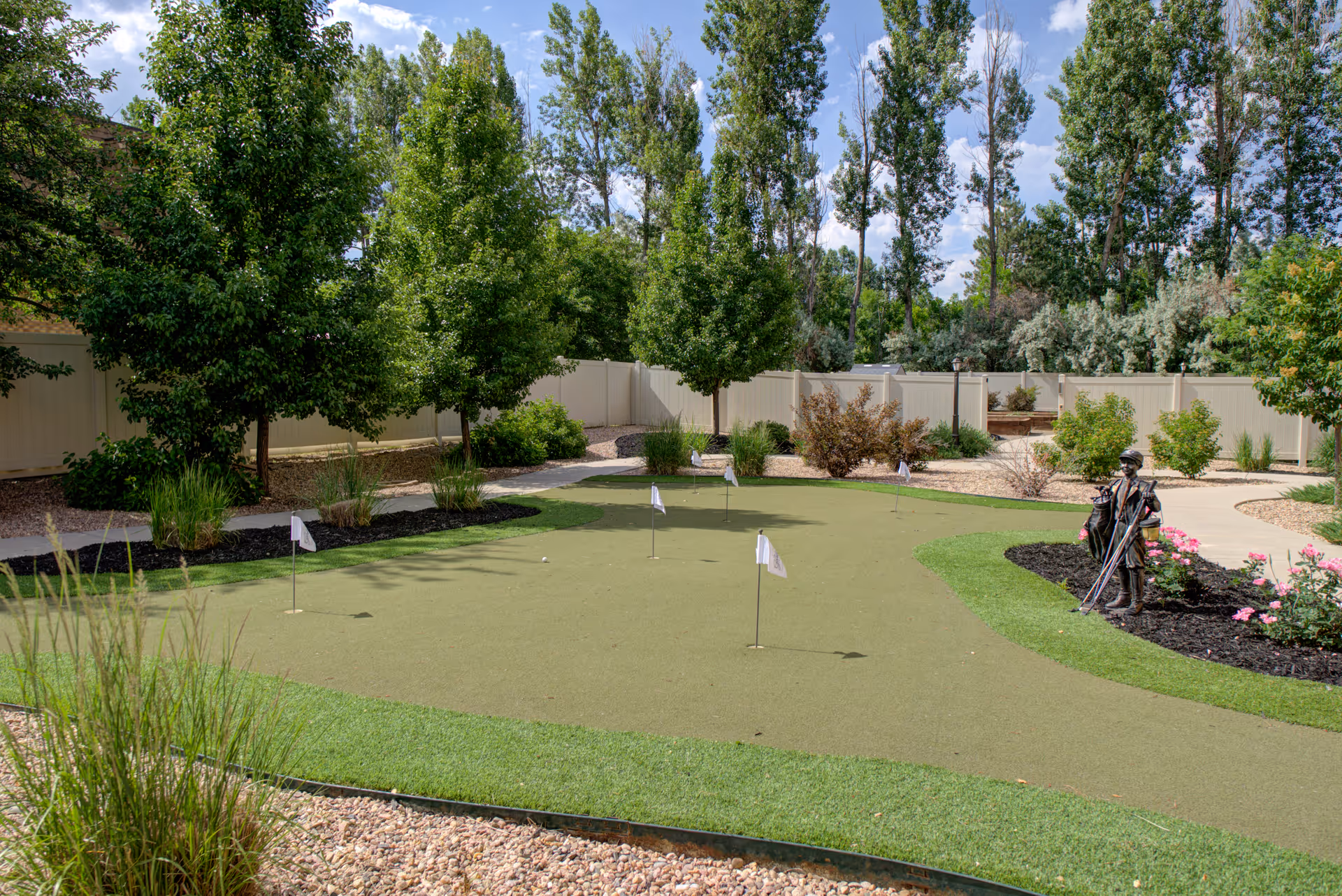 A landscaped outdoor putting green with small flags, surrounding trees, pathways, and a statue of a golfer in a fenced courtyard.