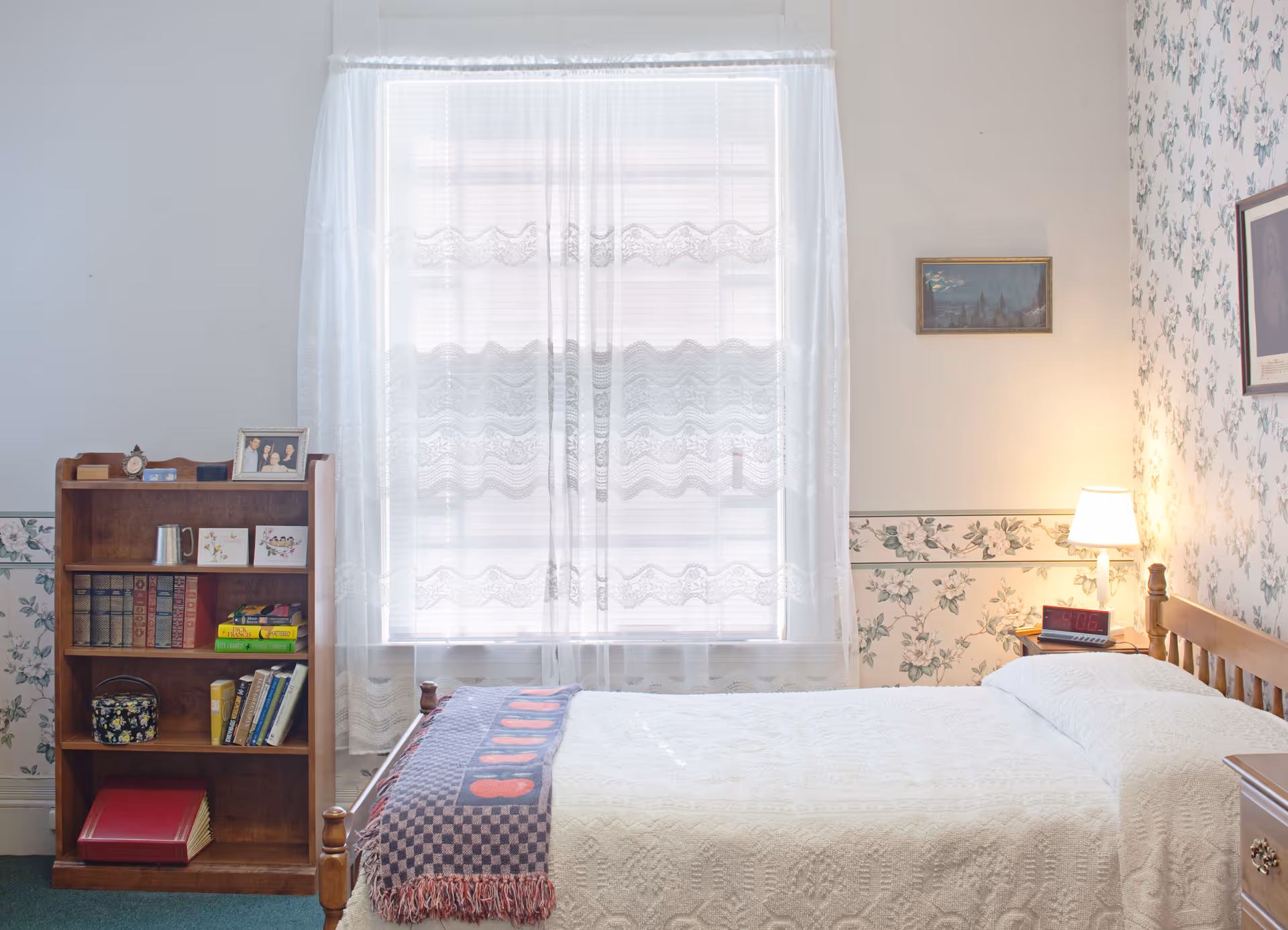 A cozy bedroom with a single wooden bed covered in a white quilt and a patterned throw blanket. To the left of the bed is a wooden bookshelf filled with books, a photo frame, and decorative items. A window with sheer white curtains lets in natural light. The walls have floral wallpaper and there is a small bedside table with a lamp and a digital clock.