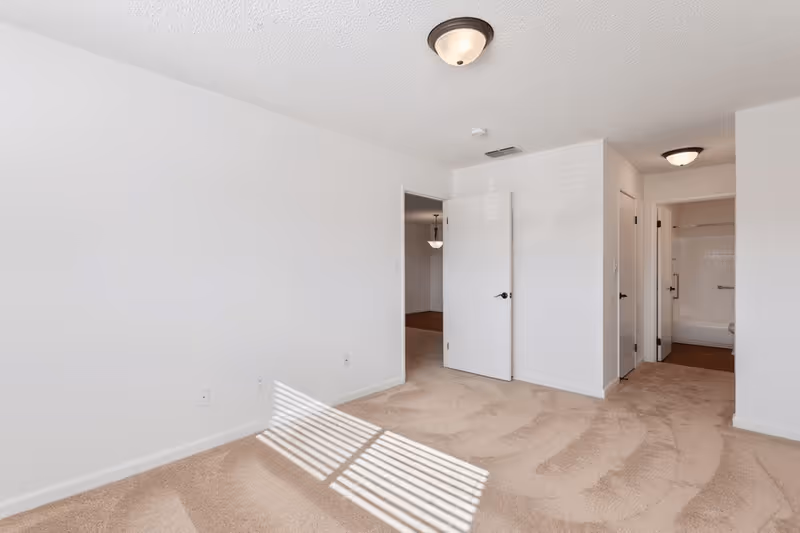 Empty room with beige carpet and white walls, featuring an open door leading to another room and a hallway with a bathroom visible at the end. Ceiling lights are mounted on the white textured ceiling.
