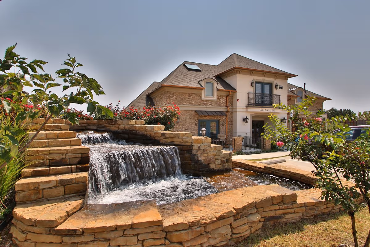 Tiered stone waterfall and pond with landscaping in front of a two-story residential building with a small balcony.