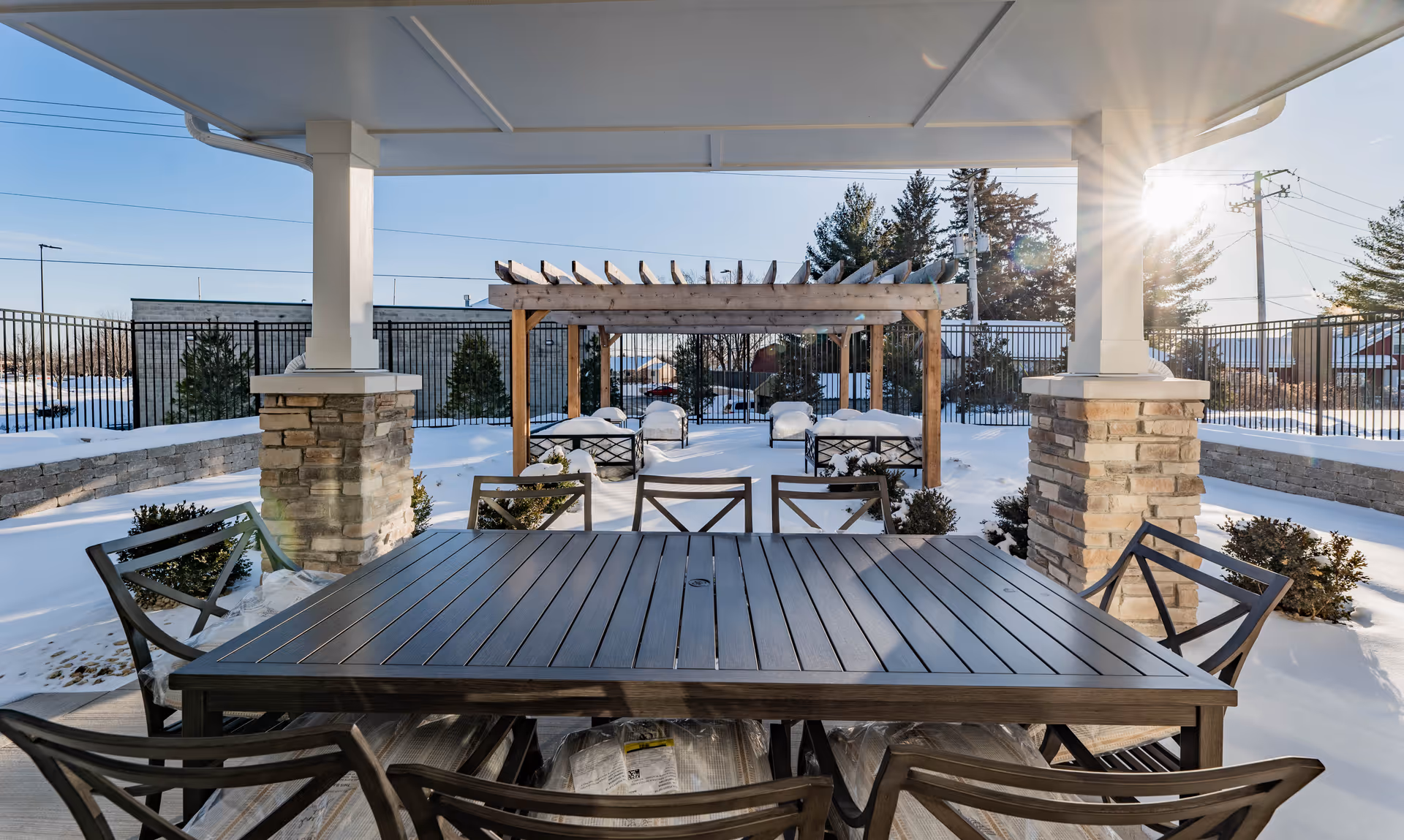 Outdoor patio area covered in snow with a large wooden table and chairs under a roofed structure. In the background, there is a wooden pergola with additional seating and snow-covered ground, surrounded by a black metal fence and some trees.