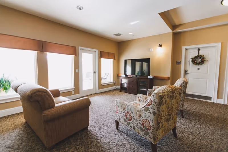 A cozy senior living room with a brown sofa and two floral armchairs arranged around a TV on a wooden stand. The room has beige walls, carpeted floor, three large windows with brown valances, and a white door decorated with a wreath.
