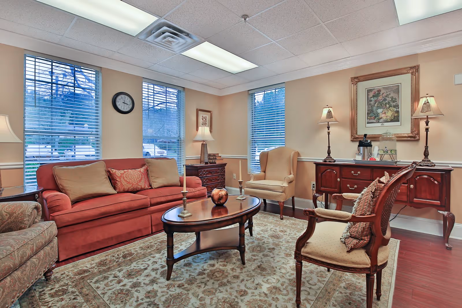 A warmly furnished living room with a red sofa, armchairs, a wooden coffee table, lamps, and windows.