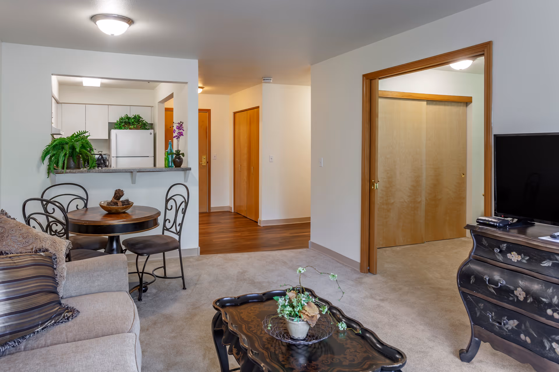 Interior view of a senior living facility apartment showing a living room with a beige sofa and decorative pillows, a black ornate coffee table with a potted plant, a round dining table with four chairs, and a kitchen area with white cabinets and a refrigerator. There is a hallway with wooden doors and an open doorway leading to another room with sliding wooden closet doors. A TV is placed on a black floral-painted dresser.