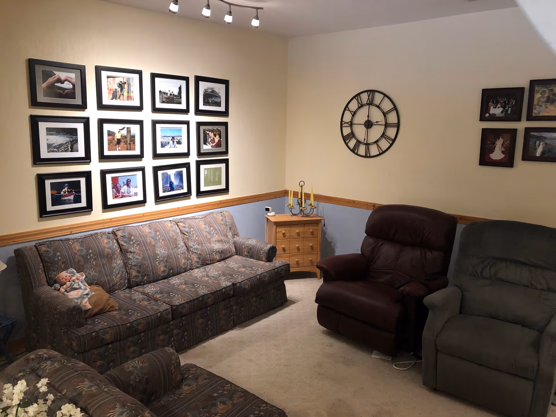A cozy living room with a floral patterned sofa and armchair, a brown leather recliner, and a green recliner. The walls are decorated with multiple framed photographs and a large black wall clock. A small wooden side table with a candelabra and a small device sits in the corner. The room is softly lit by a ceiling light fixture.