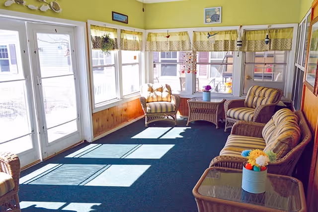 A bright sunroom with large windows and glass doors letting in natural light. The room features wicker furniture with striped cushions, including a sofa, two armchairs, and a glass-top coffee table with a small vase of colorful flowers. The walls are painted light green with wood paneling on the lower half, and there are yellow valances on the windows.