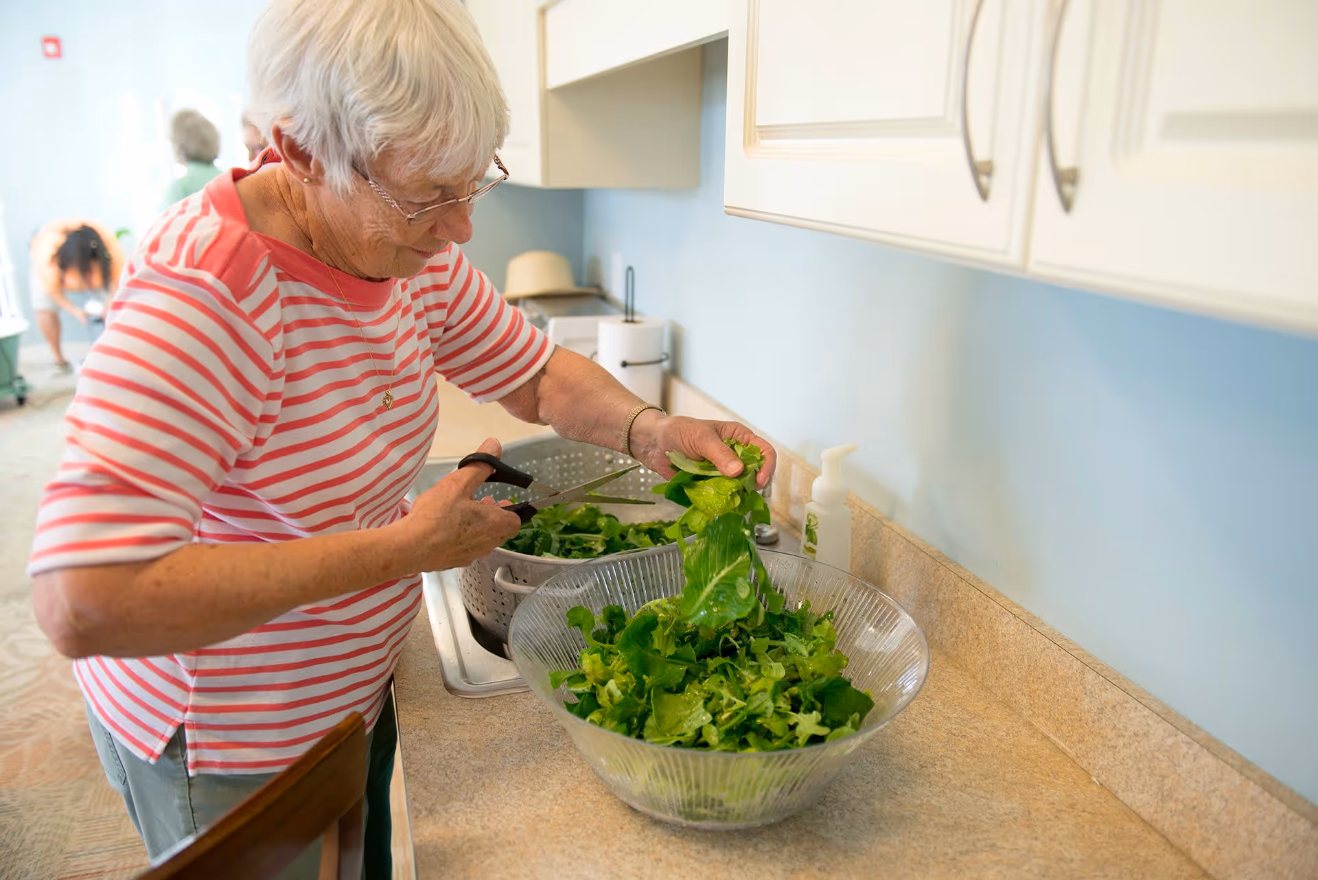 An elderly woman with white hair and glasses wearing a red and white striped shirt is cutting leafy greens over a kitchen counter with a large glass bowl and a colander nearby. The kitchen has light blue walls and white cabinets.