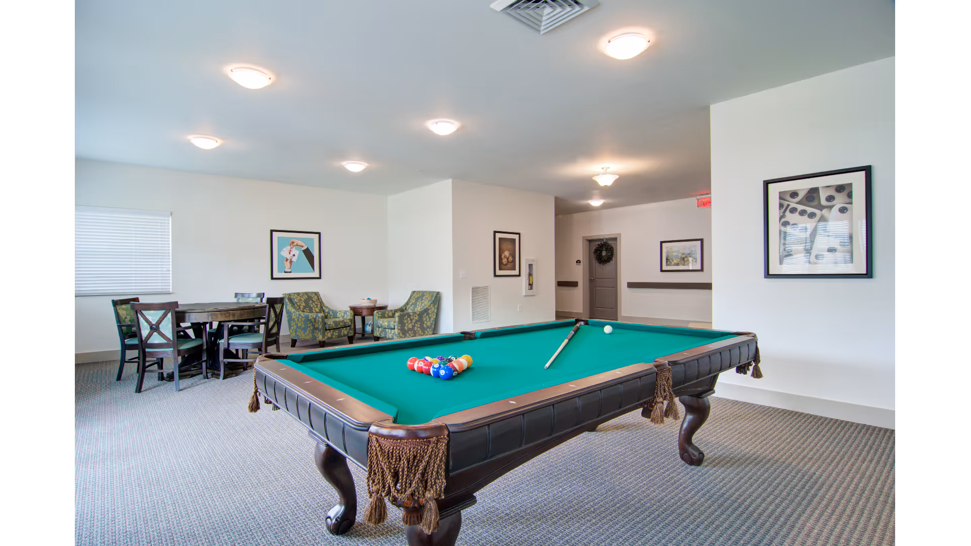 A recreational room with a green felt pool table in the center, a cue stick resting on it, and a rack of billiard balls arranged for a game. In the background, there is a round table with four chairs and two patterned armchairs with a small round table between them. The walls are decorated with framed pictures, and the room is well-lit with ceiling lights.