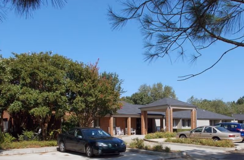 Exterior view of a single-story brick building with a covered entrance and several parked cars in front, surrounded by trees and greenery under a clear blue sky.