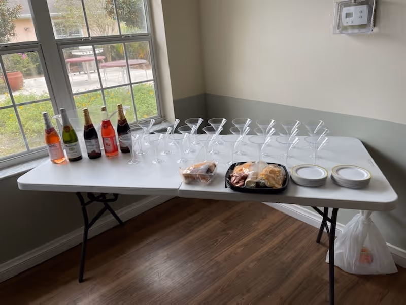 A white folding table set up indoors near a window with a view of an outdoor patio. On the table are several bottles of sparkling beverages, multiple empty martini glasses arranged in rows, a tray with assorted snacks, a small container of muffins, and two stacks of small plates. The room has wooden flooring and light-colored walls.