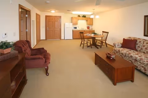 A spacious living area with beige carpet featuring a floral patterned sofa with a red pillow, a wooden coffee table, a burgundy armchair, a wooden dining table with four chairs, and a small kitchen area with wooden cabinets, a white refrigerator, microwave, and stove. The room has a ceiling fan and a wooden front door.