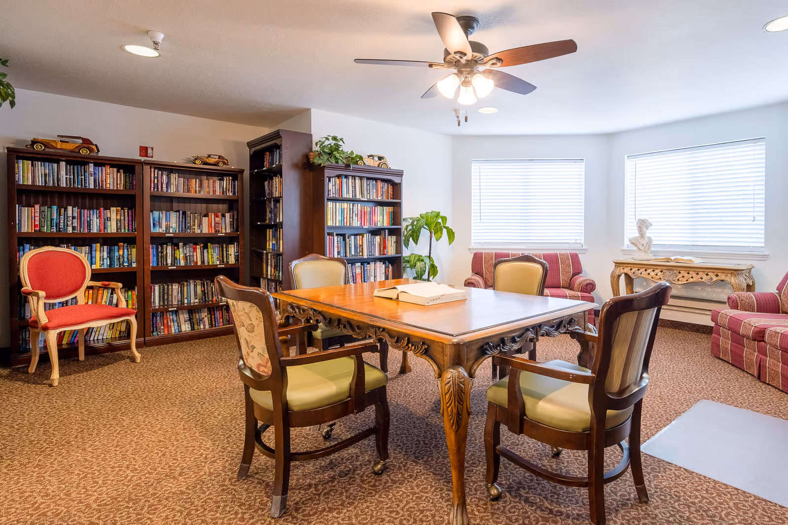 A cozy reading or common room with a wooden table surrounded by four chairs, two bookshelves filled with books, two red upholstered armchairs, a small wooden console table with a bust sculpture, and two windows with blinds letting in natural light. The room has a ceiling fan with lights and a patterned carpet.