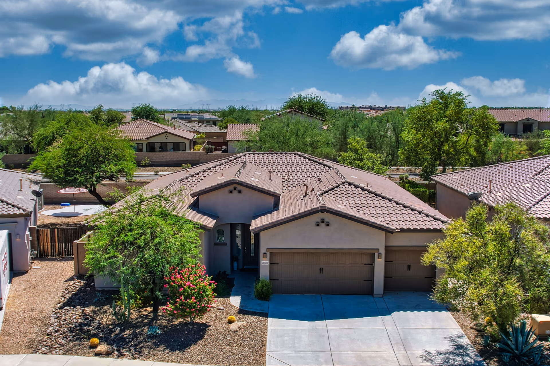 Single-story stucco house with a tiled roof, three-car garage, and desert landscaping in a suburban neighborhood under a blue sky.