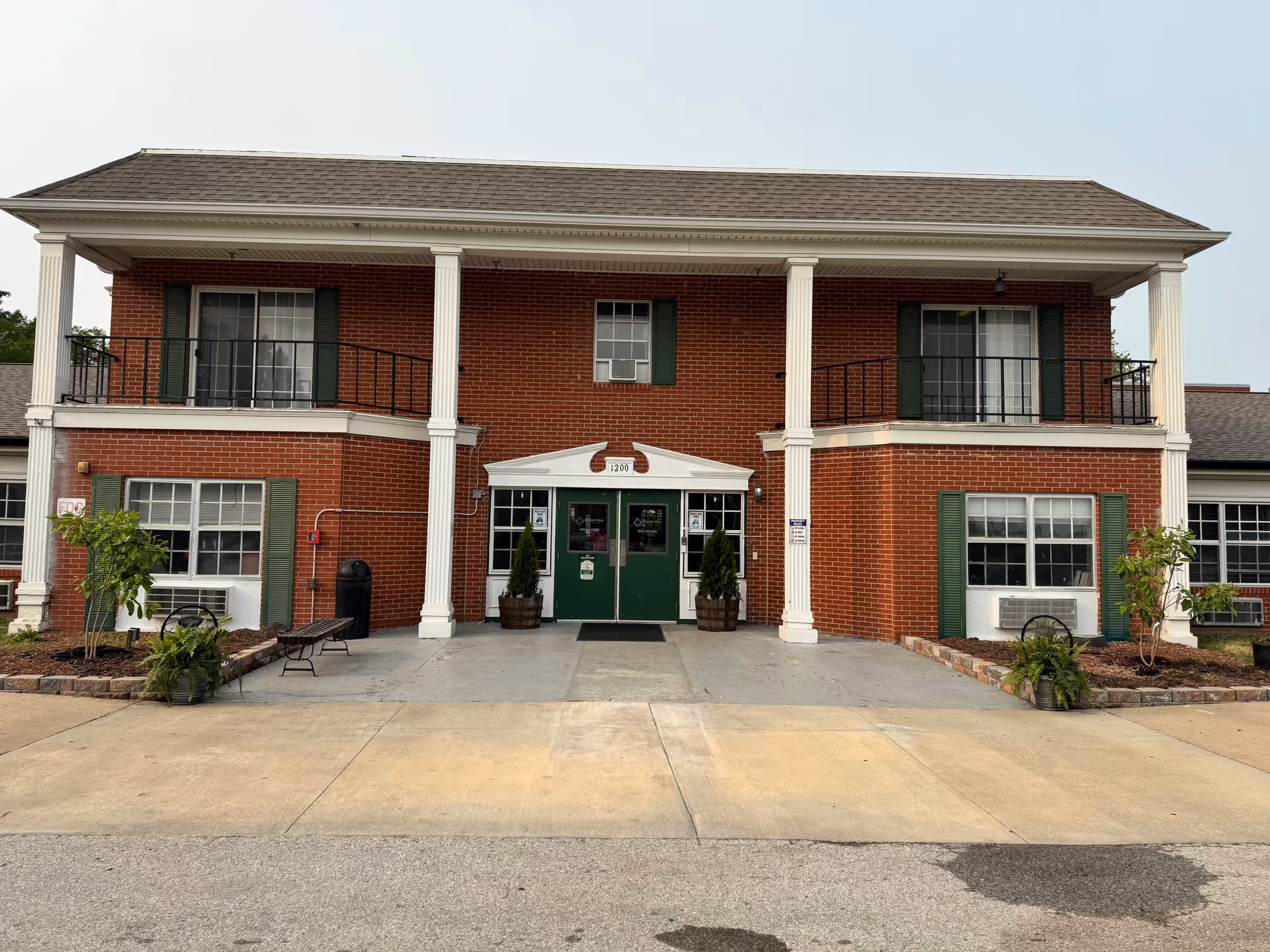 Brick two-story senior living building with white columns, green double entrance doors and second-floor balconies.