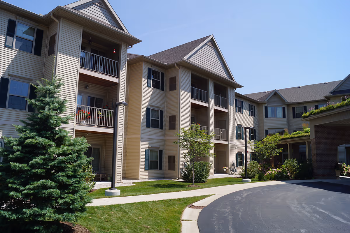 Exterior view of a multi-story beige senior living building with balconies, landscaped lawns, and a curved driveway.