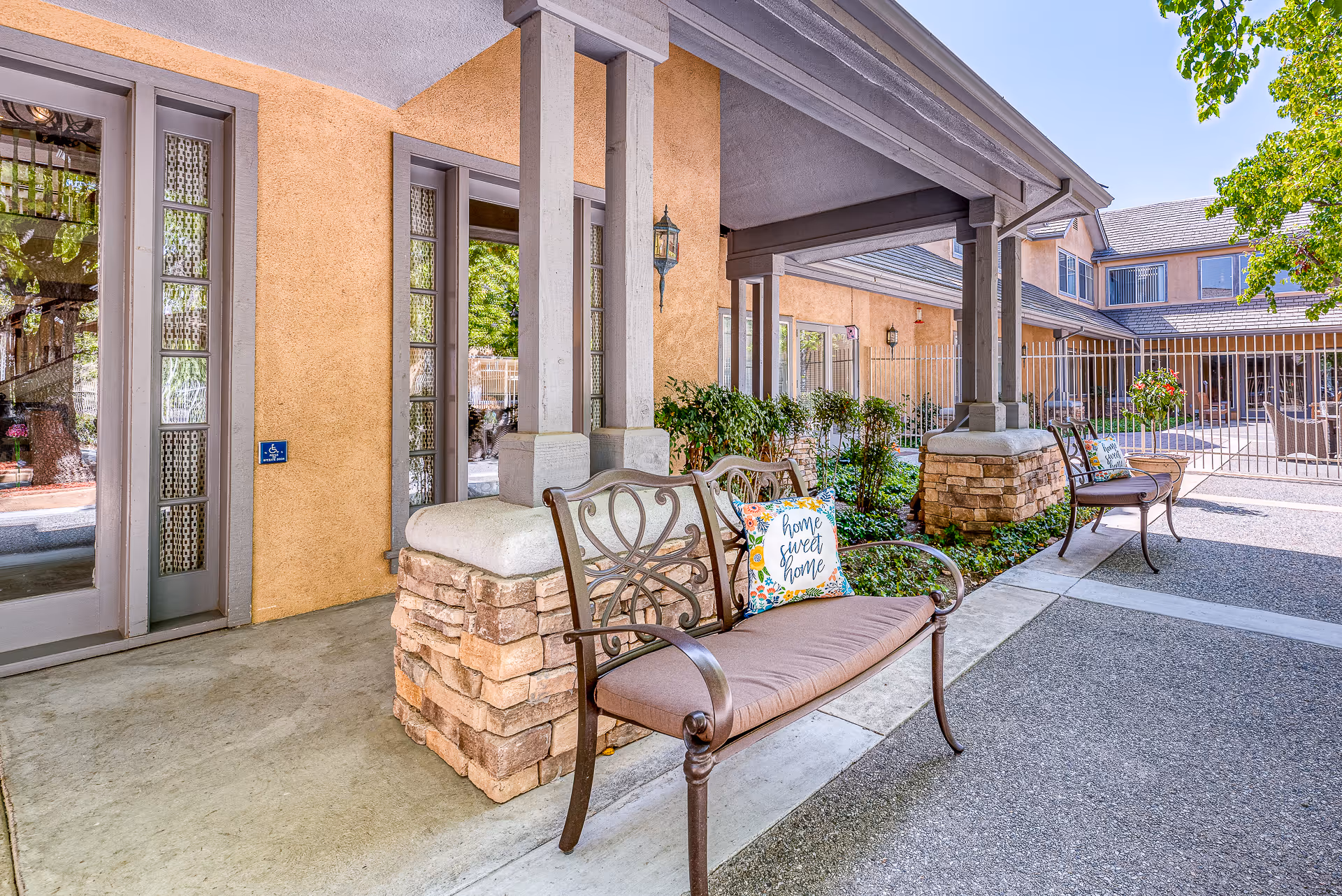 Outdoor covered seating area at Chino Hills Senior Living with metal benches featuring cushions and decorative pillows that say 'home sweet home'. The area has stone pillars, beige walls, large windows, and a gated courtyard with plants and trees under a clear blue sky.