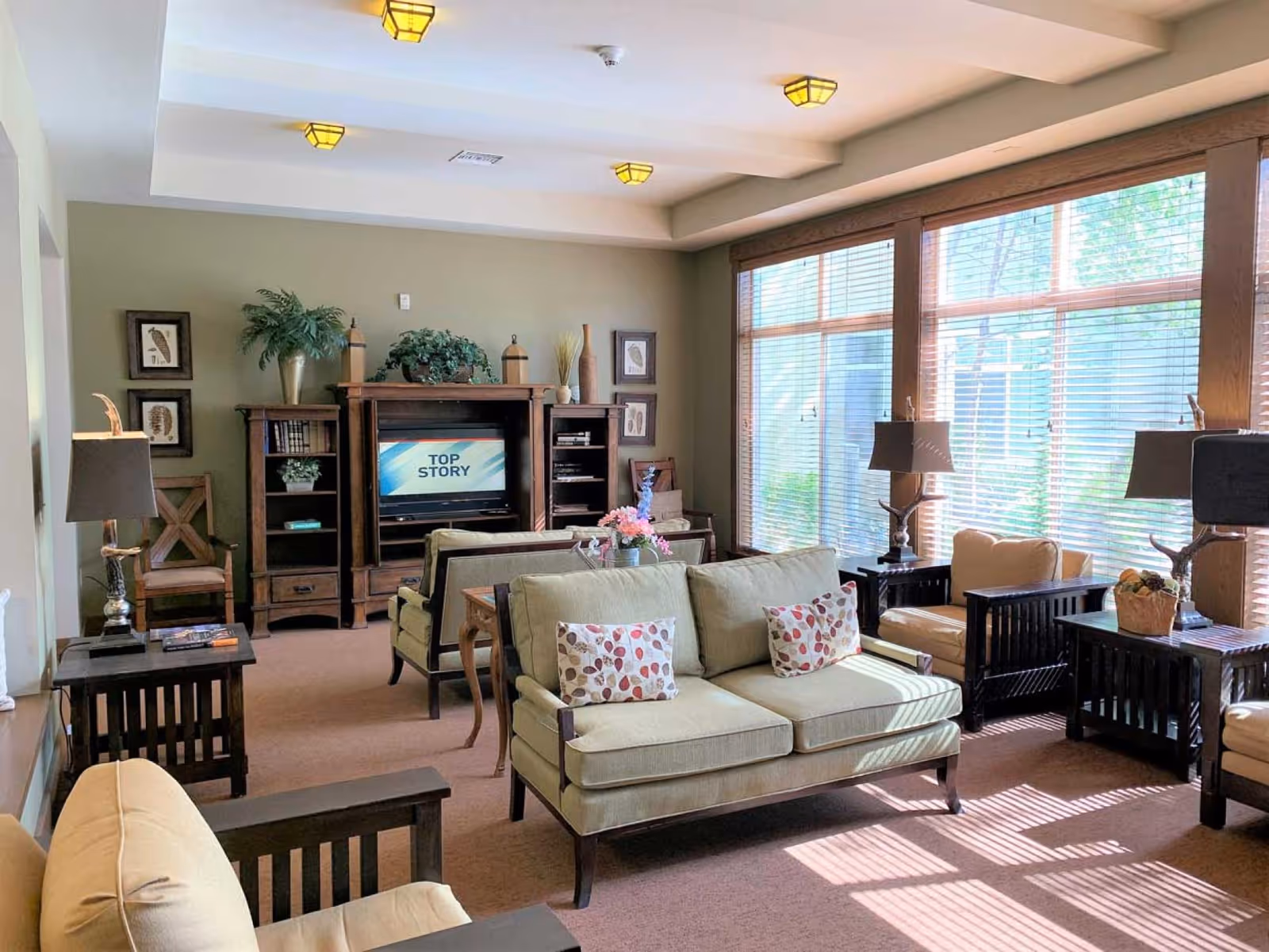 A bright and cozy living room with beige sofas and armchairs featuring wooden frames and patterned cushions. The room has large windows with wooden blinds allowing natural light to fill the space. There are two dark wooden side tables with matching lamps, a wooden entertainment center with a TV displaying the text 'TOP STORY', and decorative plants and framed artwork on the walls.