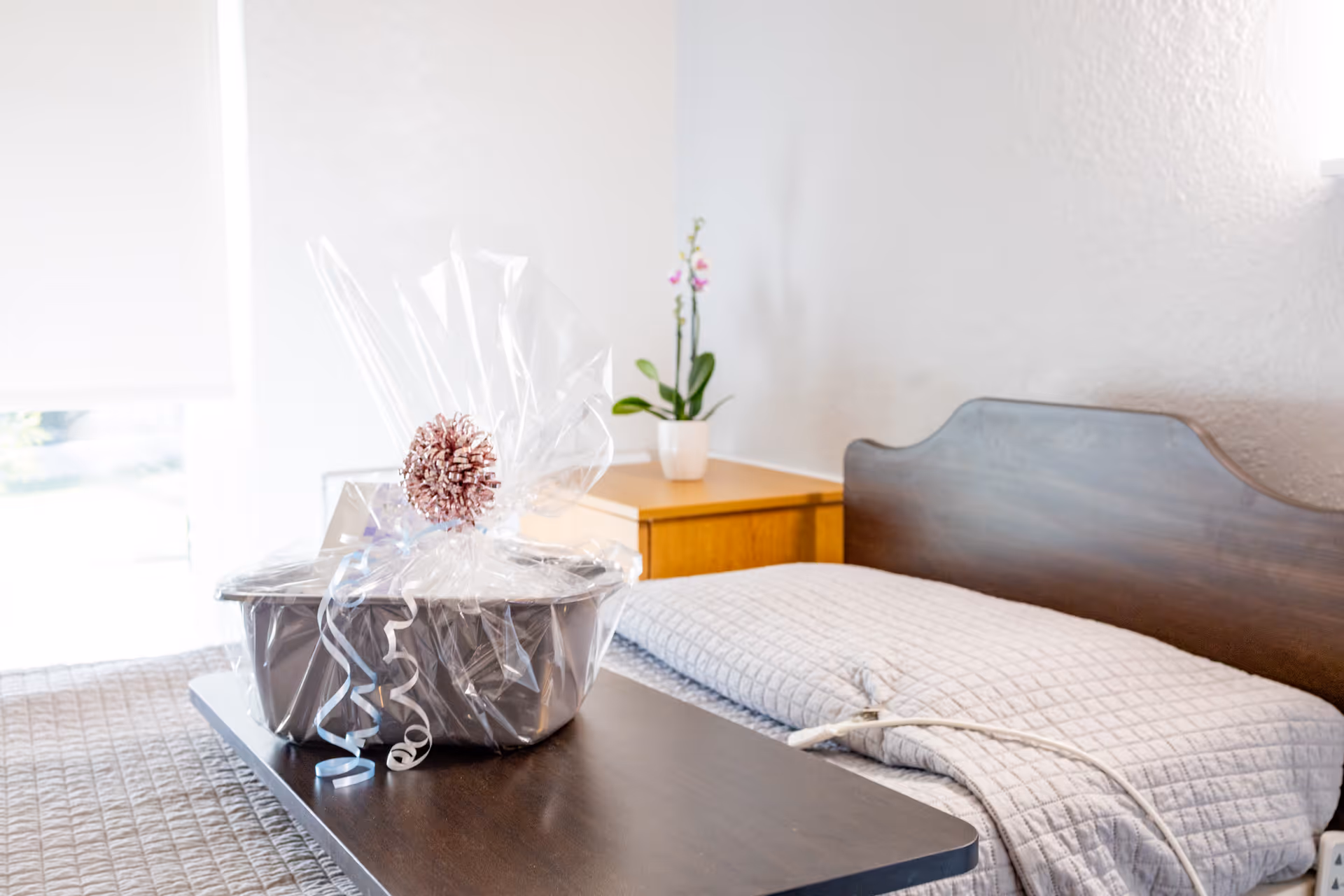 A neatly made bed with a wooden headboard in a bright room. On the bed is a dark wooden tray holding a gift basket wrapped in clear plastic with a decorative bow. A small wooden nightstand with a potted orchid plant is beside the bed.