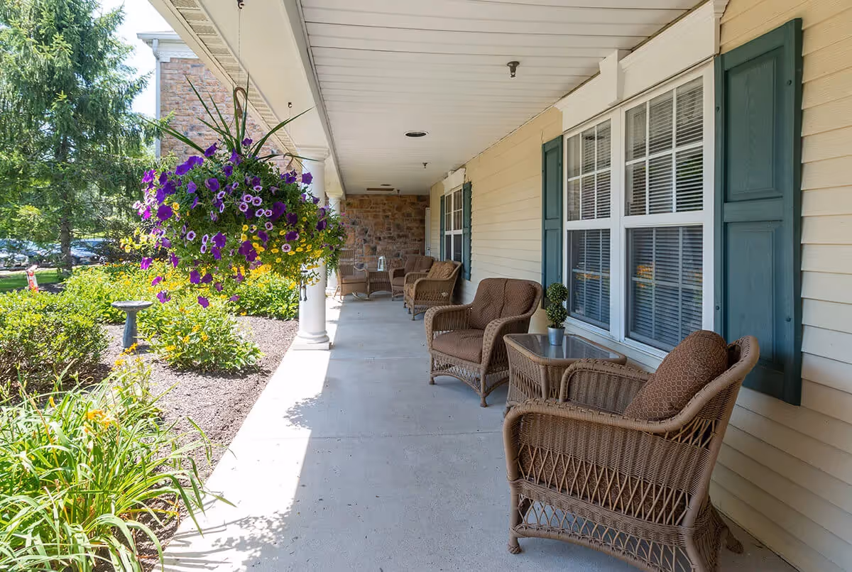 A covered outdoor porch area with wicker chairs and small tables arranged along the wall of a building. Hanging baskets with purple and yellow flowers are suspended from the ceiling. There are green shrubs and flowering plants along the edge of the porch, and a birdbath is visible in the garden area.