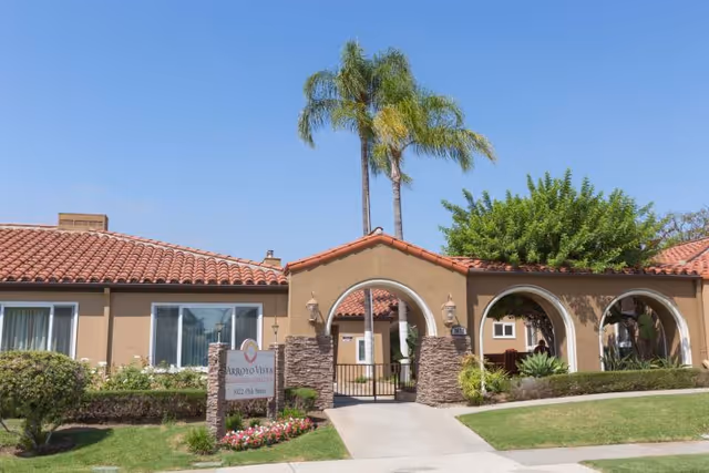 Exterior view of Arroyo Vista Nursing Center featuring a beige building with a red tile roof, arched entryways, palm trees, and well-maintained landscaping including bushes and flowers under a clear blue sky.