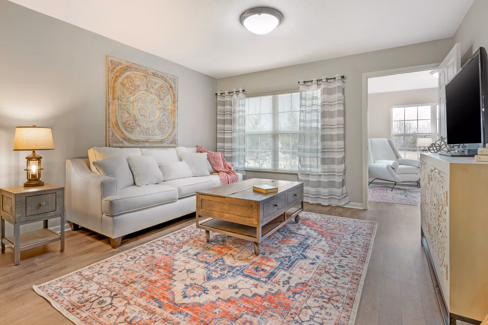 A bright living room with a white sofa adorned with multiple cushions and a pink throw blanket. A wooden coffee table with drawers sits on a colorful patterned rug. To the left, there is a wooden side table with a lamp. Large windows with sheer striped curtains allow natural light to fill the room. In the background, an adjacent room with a white armchair and a window is visible. A flat-screen TV is mounted on a decorative cabinet to the right.