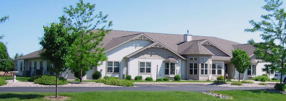 Exterior view of a single-story building with beige siding and a brown roof, surrounded by green grass, trees, and shrubs under a clear blue sky.