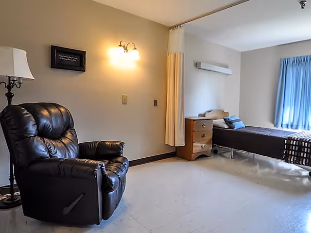 A simple, clean room with a black leather recliner chair on the left, a floor lamp beside it, and a wall-mounted light fixture above. On the right side, there is a single bed with a brown blanket and blue pillows, a wooden nightstand with drawers, and a window with blue curtains letting in natural light.