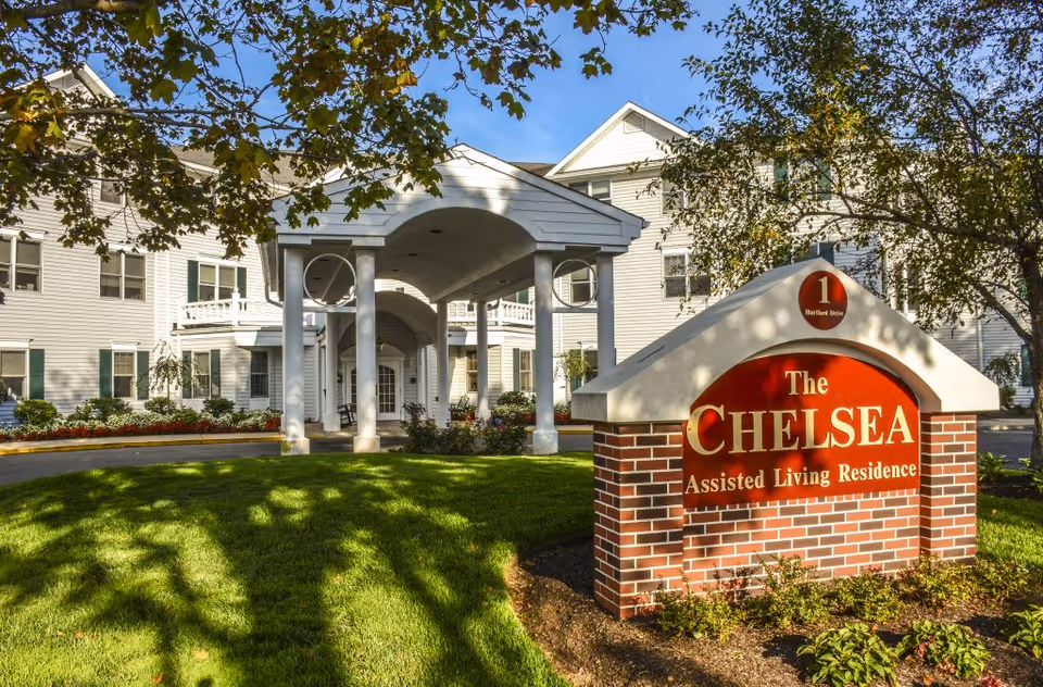 Exterior view of The Chelsea Assisted Living Residence building with a covered entrance supported by white columns, surrounded by green grass, trees, and landscaping under a clear blue sky.
