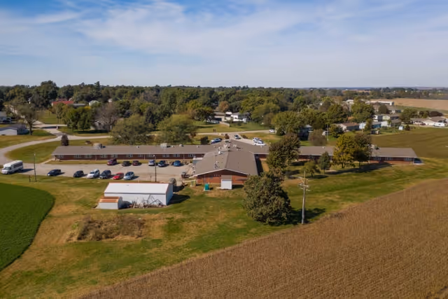 Aerial view of Falls City Nursing and Rehabilitation Center, a single-story building with a large parking lot and several cars parked. The building is surrounded by green lawns, trees, and farmland under a partly cloudy sky.