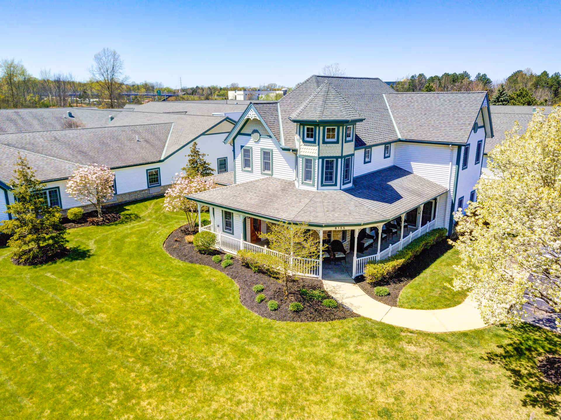 A large, white senior living facility building with green trim and a wraparound porch surrounded by well-maintained green lawns and blooming trees under a clear blue sky.