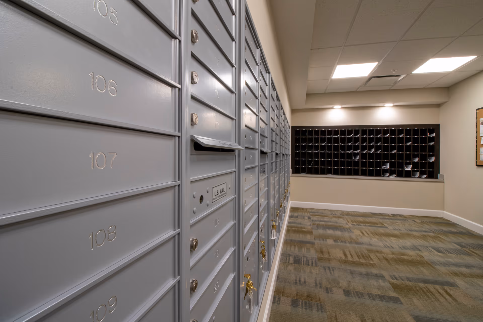 A mailroom with rows of gray metal mailboxes on the left wall and a large set of black cubbyholes on the far wall. The room has a carpeted floor with a patterned design and a ceiling with recessed lighting.