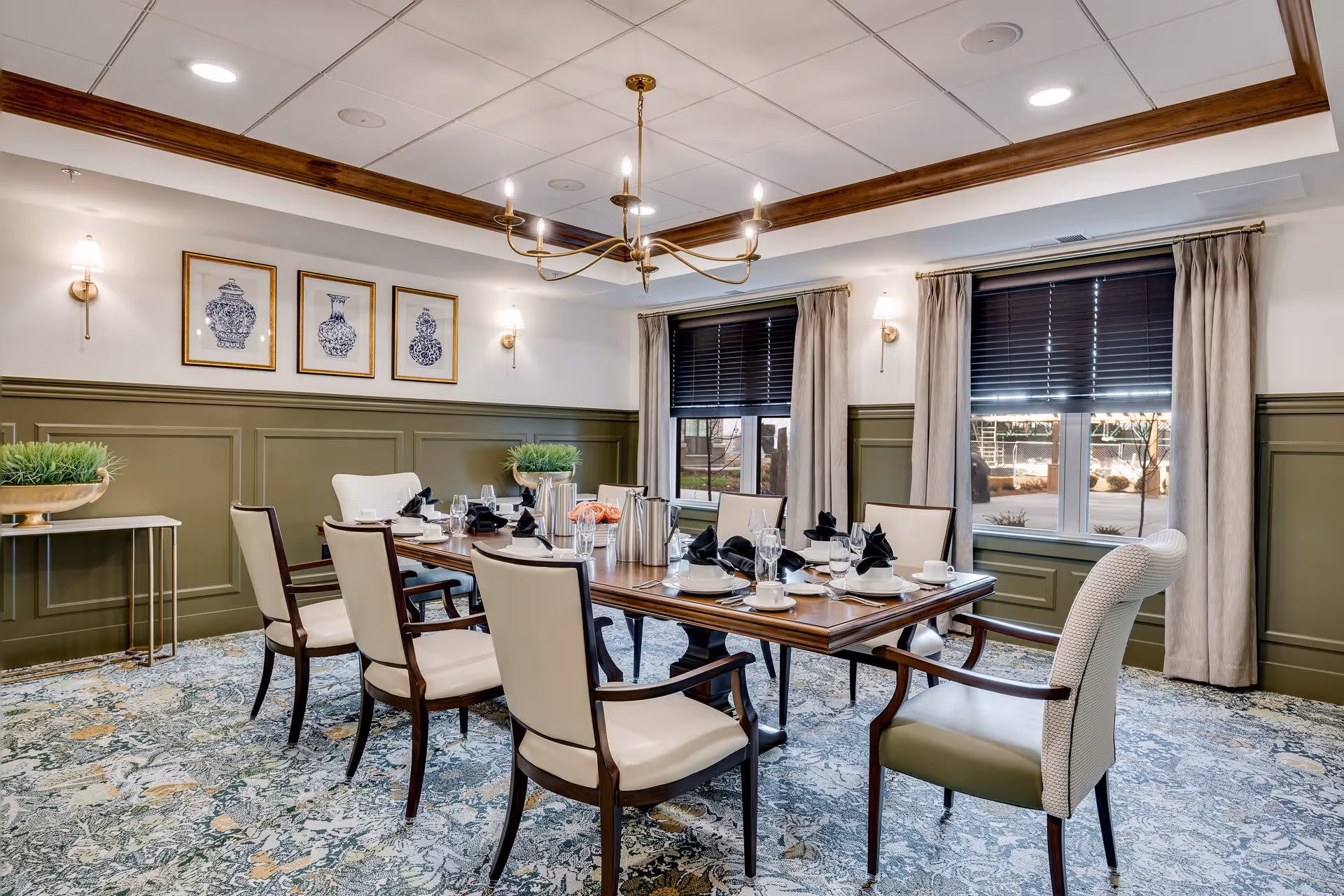 A formal dining room with a wooden table set for eight people. The table is arranged with white plates, cups, glasses, and black napkins. The room has green wainscoting on the walls, beige curtains, and two windows with dark blinds. There are three framed blue and white vase prints on the wall, a chandelier hanging from the ceiling, and two wall sconces providing light. The floor is covered with a patterned carpet in shades of blue, green, and beige.