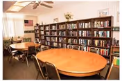 A room with multiple wooden tables and chairs arranged for seating. Along one wall, there is a large bookshelf filled with books. The room has a ceiling fan, vertical blinds on the windows, and an American flag hanging on the wall.