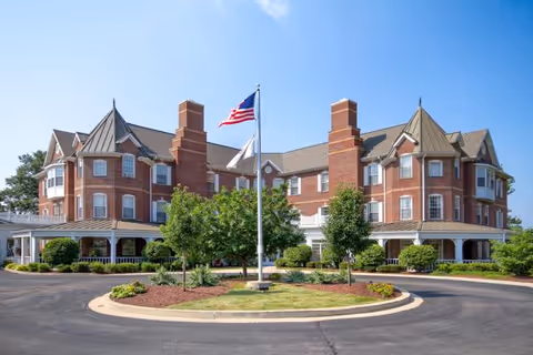 Front exterior view of a large, multi-story brick building with multiple chimneys and peaked roofs. The building is surrounded by well-maintained landscaping including bushes and trees, with an American flag flying on a flagpole in the center of a circular driveway.