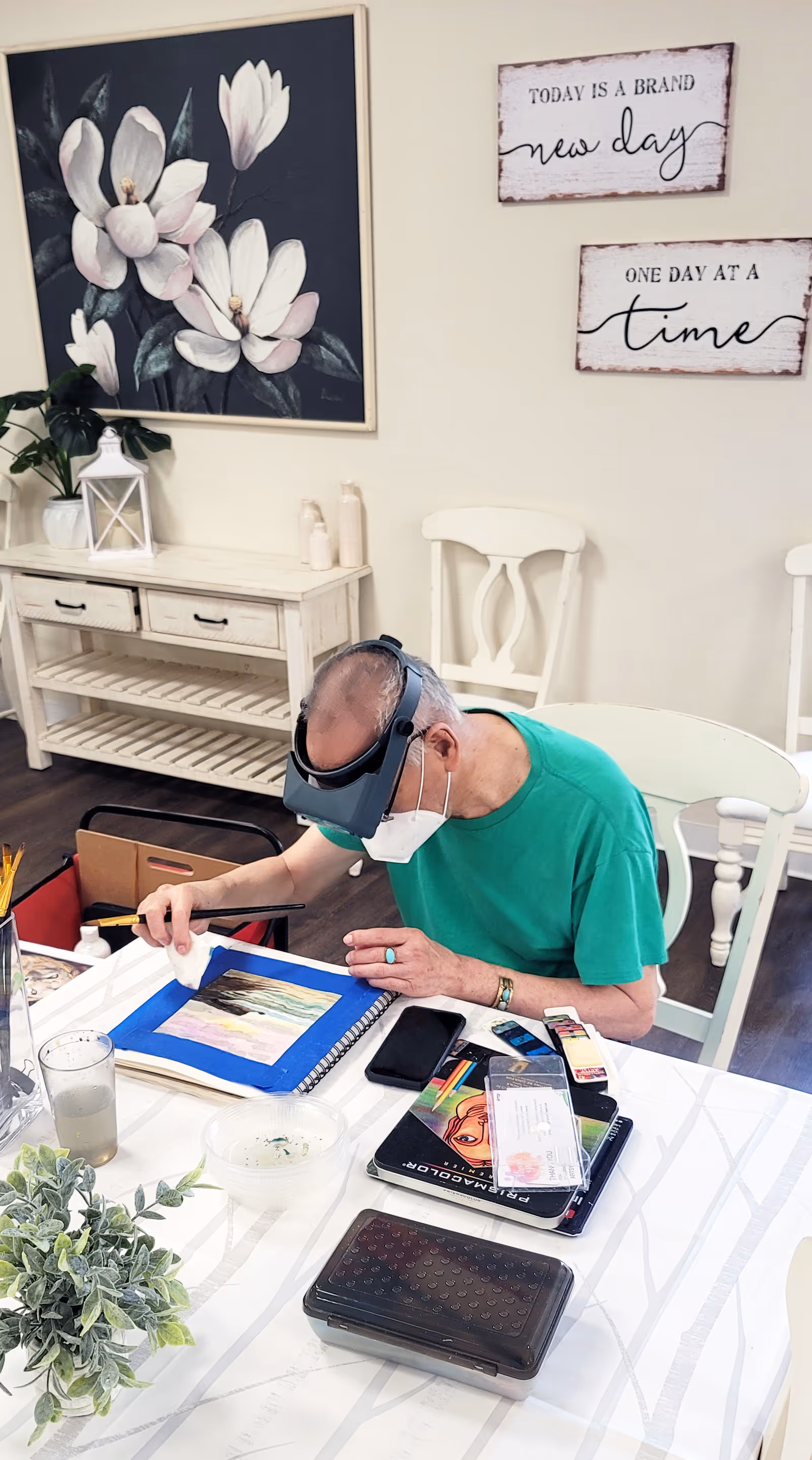 An elderly man wearing a green shirt and a face mask is seated at a table engaged in painting or drawing. He is using a brush on a piece of paper taped to a blue mat. The table has various art supplies, a glass of water, and a small plant. Behind him is a white wooden console table with decorative items and two white chairs. On the wall are three framed artworks, including a large floral painting and two signs with motivational quotes: 'TODAY IS A BRAND new day' and 'ONE DAY AT A time'.
