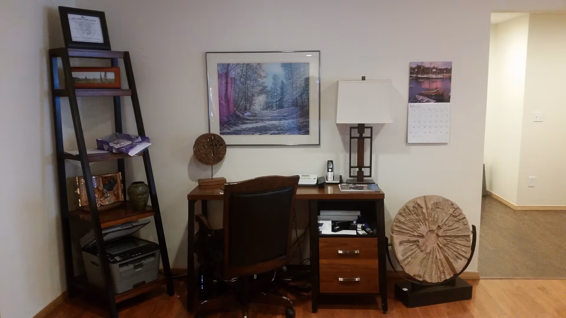 A small office corner with a wooden desk, a black office chair, a table lamp, a cordless phone, and some papers. To the left of the desk is a dark wooden ladder-style shelf holding various items including a printer and decorative pieces. On the wall above the desk is a framed picture of a forest path, and to the right is a calendar. A large circular decorative piece is placed on the floor next to the desk.
