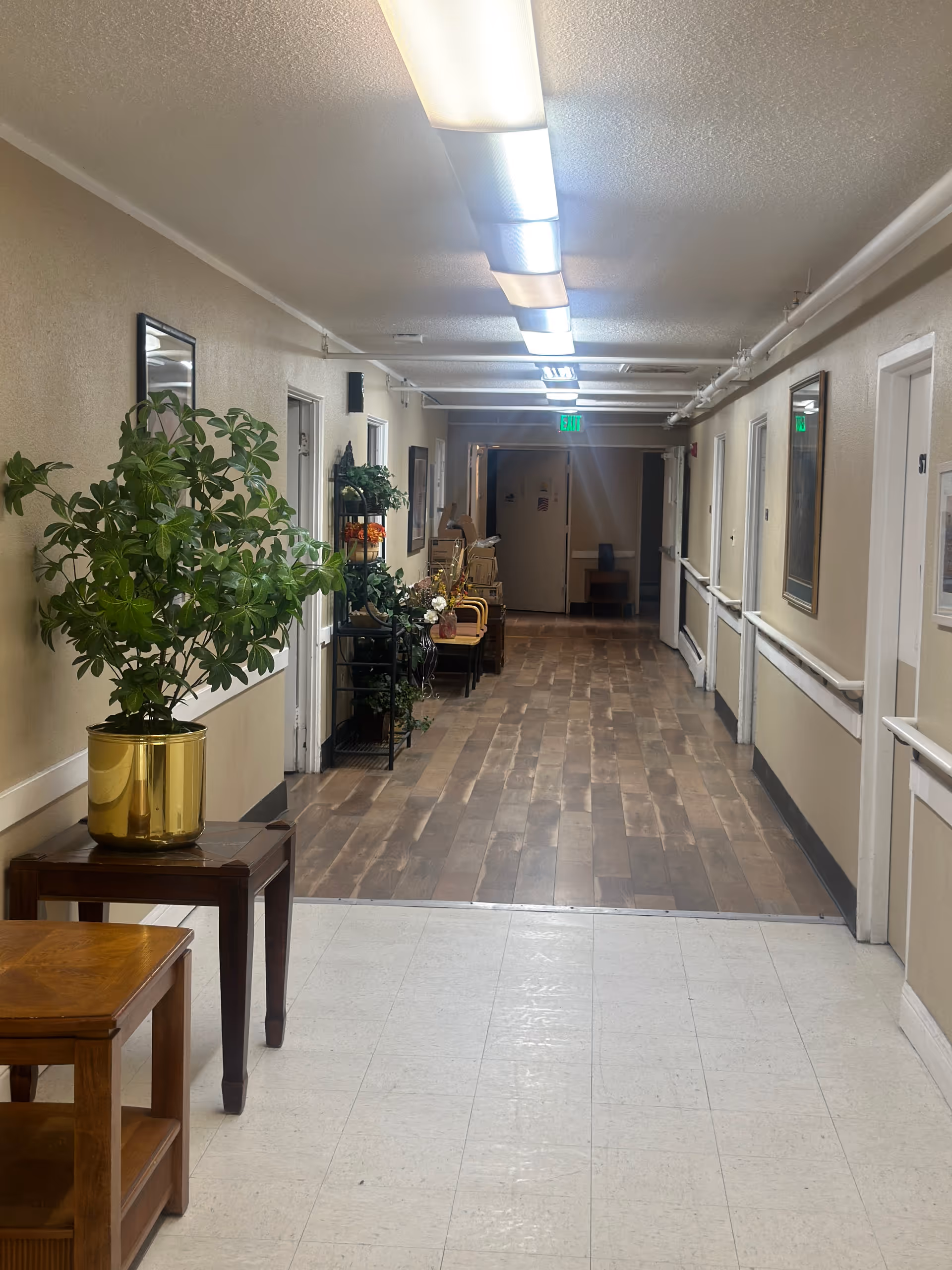Long interior hallway of an assisted living facility with potted plants, chairs, handrails and overhead lights.