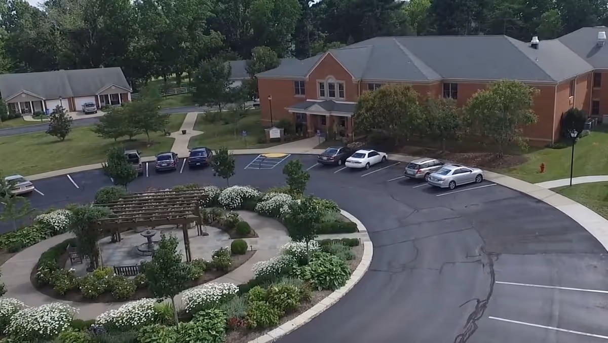 Aerial view of Wesley Village Senior Living showing a circular driveway with parked cars, a landscaped garden area with a wooden pergola and benches, and a brick building surrounded by trees and greenery.