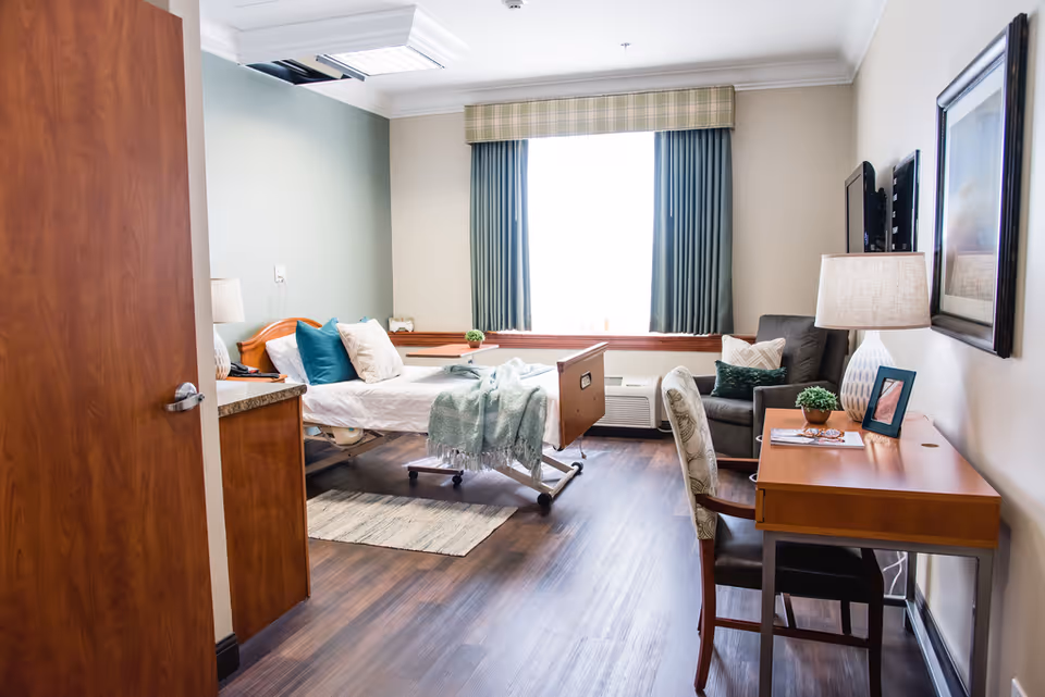 A bright senior care bedroom featuring a hospital-style bed, desk and chair, an armchair, and a window with curtains.