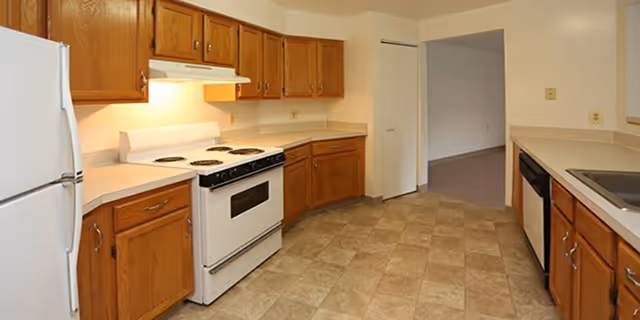 A kitchen with wooden cabinets, a white refrigerator, a white stove with an overhead vent, a dishwasher, and a sink. The floor is tiled with a beige pattern, and there is an open doorway leading to another room.