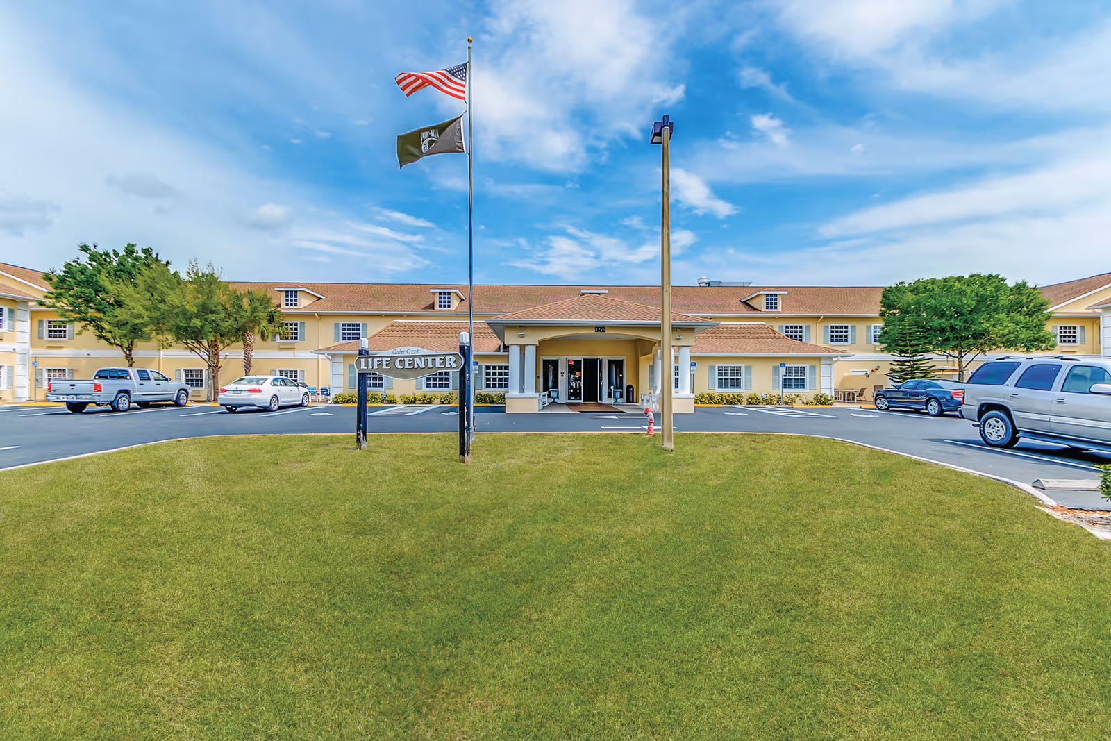 Front exterior view of Cedar Creek Assisted Living Life Center building with a green lawn in the foreground, two flagpoles with American and another flag, several parked cars, and a partly cloudy blue sky.