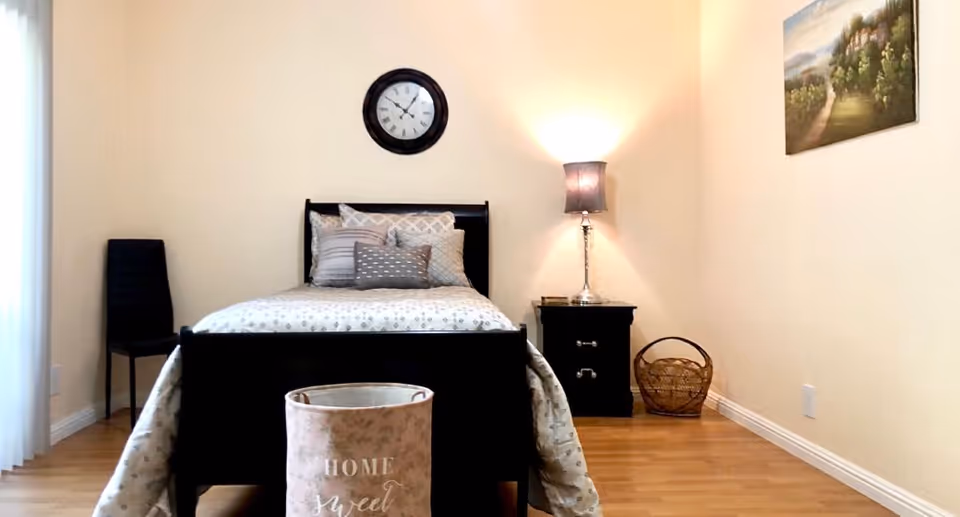 Simple furnished bedroom with a single bed centered against the wall, a nightstand and lamp to the right, a clock above the bed, and a basket on the floor.