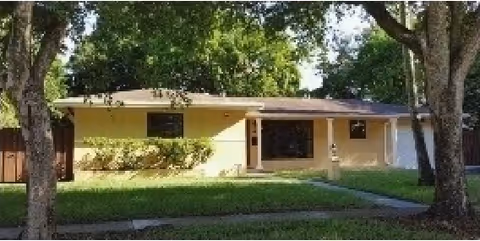 Single-story yellow house with a front porch, surrounded by green grass and trees on a sunny day.