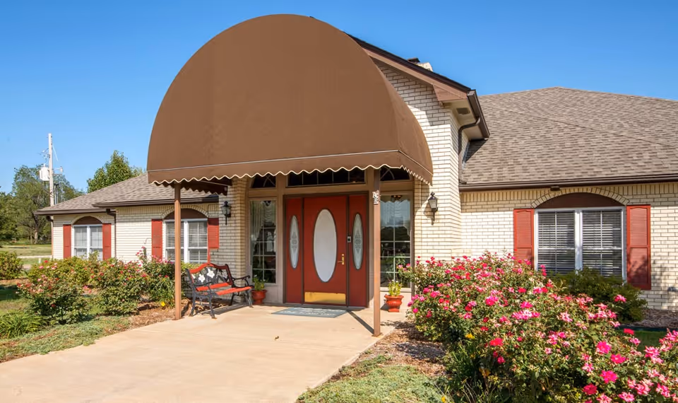 Front exterior view of a single-story brick building with a brown awning over the entrance. The entrance has double red doors with oval glass panels. There are red shutters on the windows and flower beds with pink flowers along the walkway leading to the door. A bench is placed near the entrance.