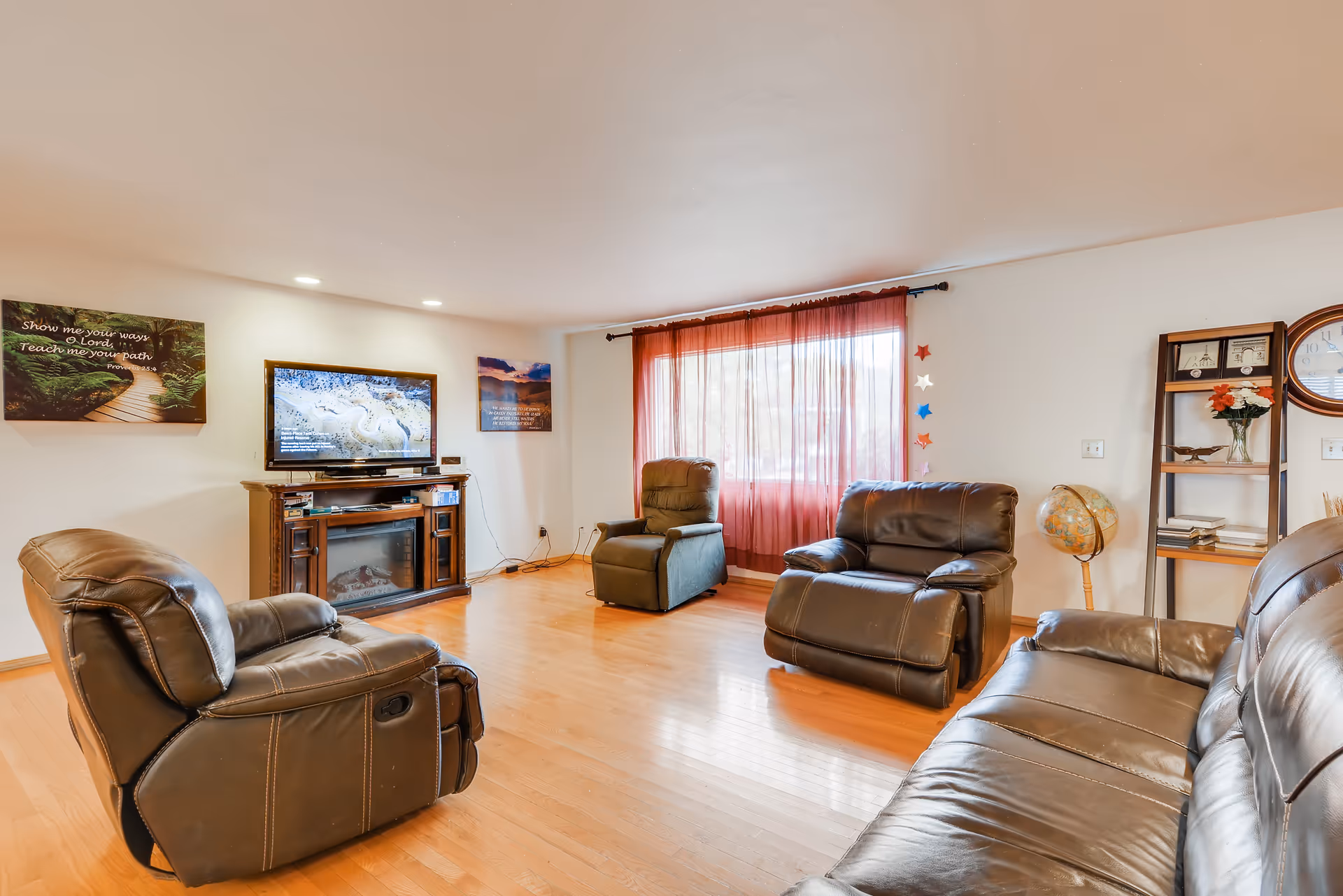 A cozy living room with four leather recliner chairs arranged around a TV mounted on a wooden stand with an electric fireplace. The room has hardwood floors, a large window with sheer red curtains, a wooden shelf with books and flowers, a globe, and wall decorations including framed quotes and a clock.
