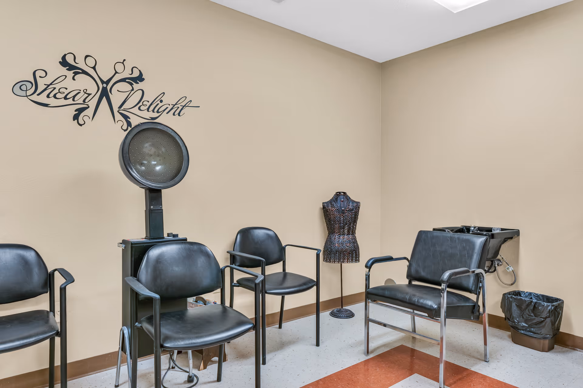 Interior of a hair salon area with four black chairs, a hair dryer, a decorative mannequin torso, and a hair washing station with a black sink and trash bin. The wall has a decorative sign that reads 'Shear Delight' with a scissors graphic.